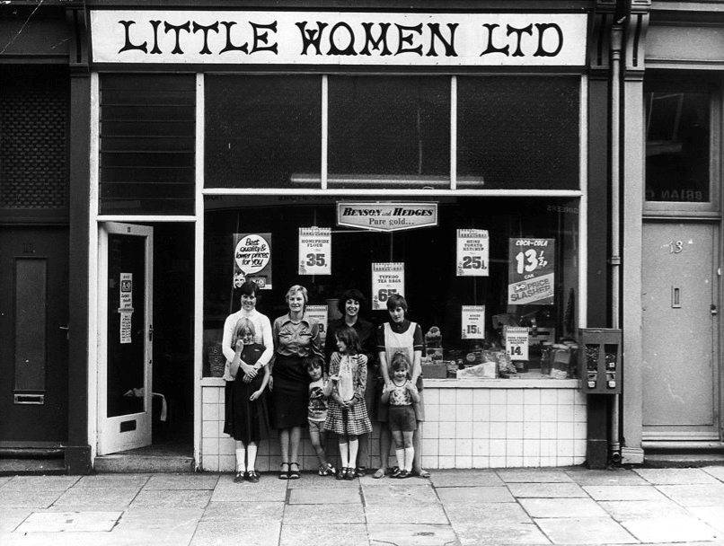 a black and white photograph showing four women standing and four children wearing 1970s clothesstanding outside a shop under a sign that says Little Women Ltd